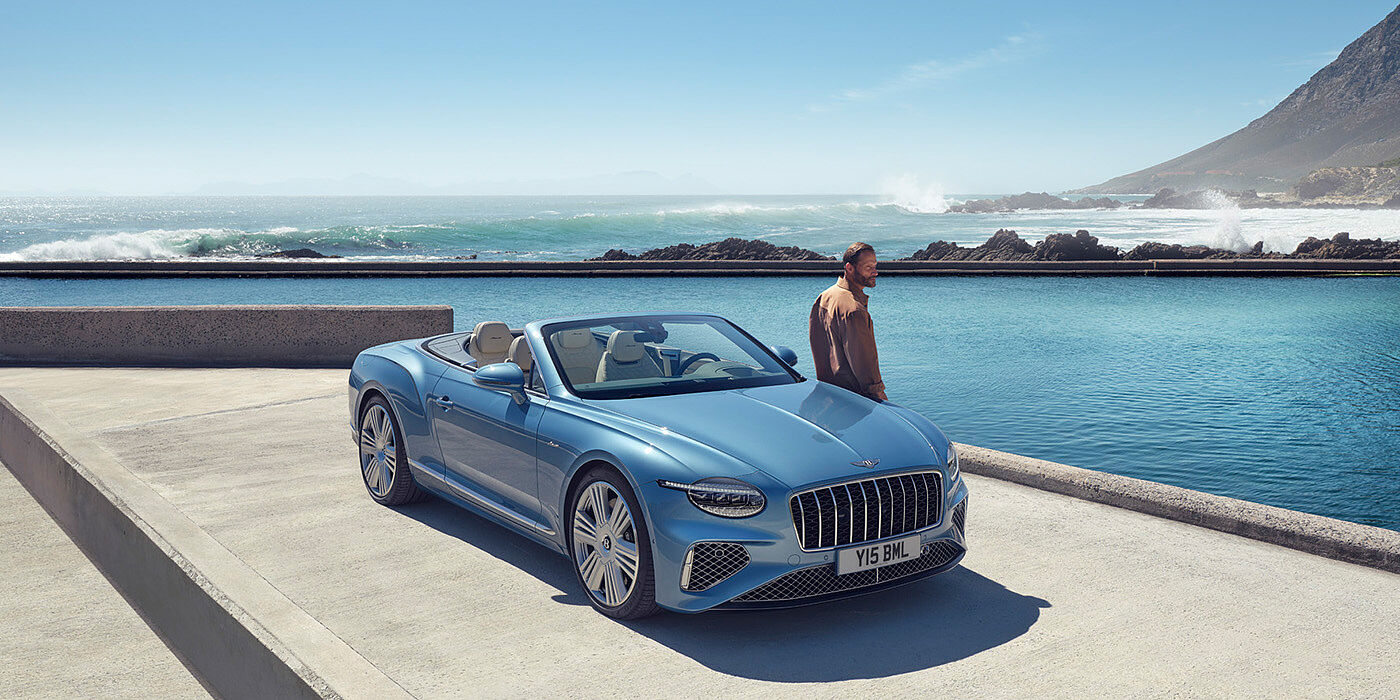 Bentley Auckland New Bentley Continental GTC Azure convertible front view in Light Windsor blue paint with 22 inch ten spoke wheels parked by the sea in the sunshine