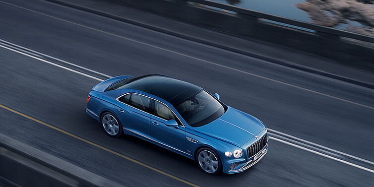 Bentley Auckland New Bentley Flying Spur Azure sedan in Light windsor blue paint by Mulliner, with 22 inch Azure wheels seen from overhead driving dynamically on a country road