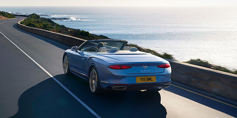 Bentley Auckland New Bentley Continental GTC Azure convertible rear view in Light Windsor blue paint with 22 inch ten spoke wheels driving dynamically on an ocean road in the sunshine