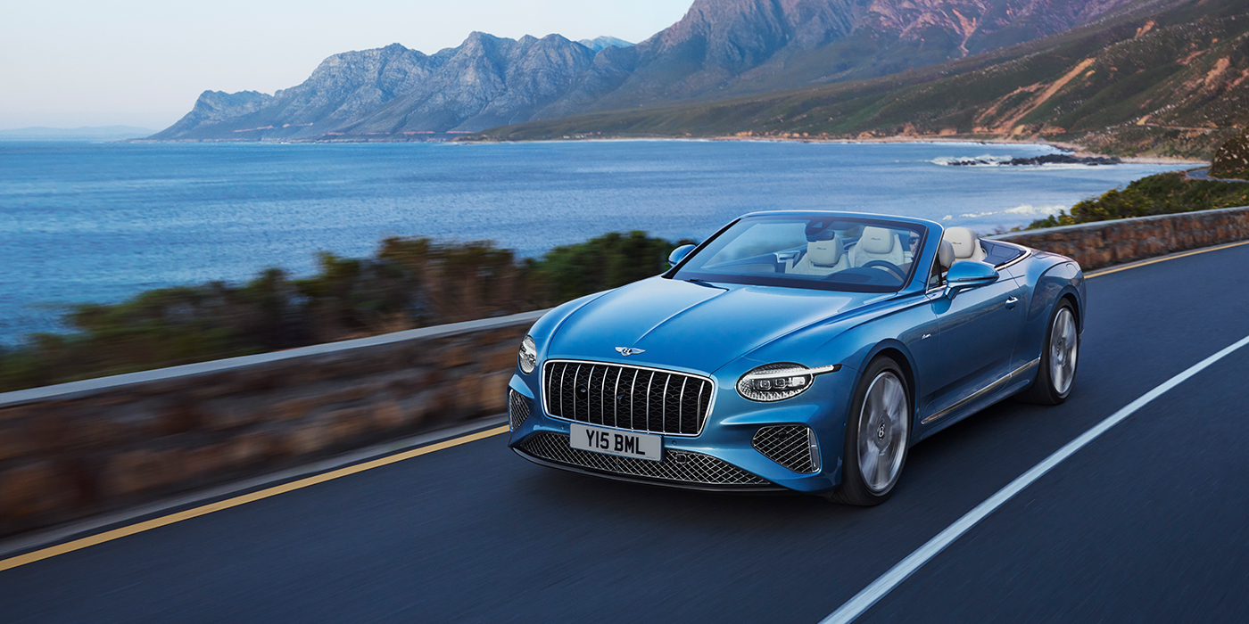 Bentley Auckland New Bentley Continental GTC Azure convertible in Light Windsor blue paint with 22 inch Azure wheels and chrome grill, driving dynamically by the sea